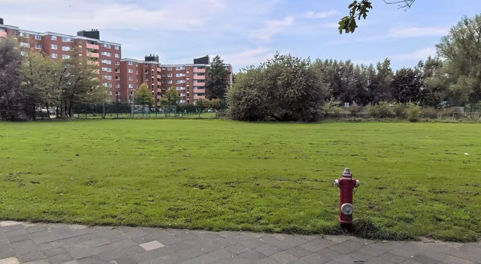 Eine große, flache Rasenfläche liegt im Vordergrund, rechts steht ein roter Hydrant am Rand eines gepflasterten Weges. Im Hintergrund sind mehrere mehrstöckige Wohngebäude aus rotem Backstein sowie Bäume und Sträucher zu sehen.